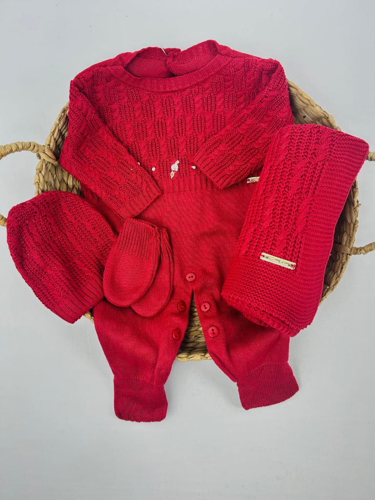 Conjunto Saída Maternidade Vera em tricô vermelho com macacão, manta, gorro e luvas, dispostos sobre cesta de palha